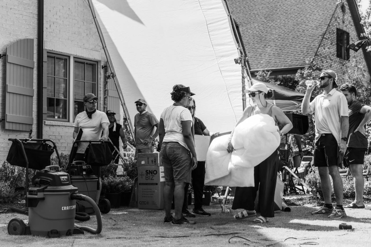 A black and white photo of a film set. A group of people stand on a grassy area, some holding equipment like boxes and a large light diffuser. A vacuum cleaner is visible on the ground. A building with a window and shutters is in the background.