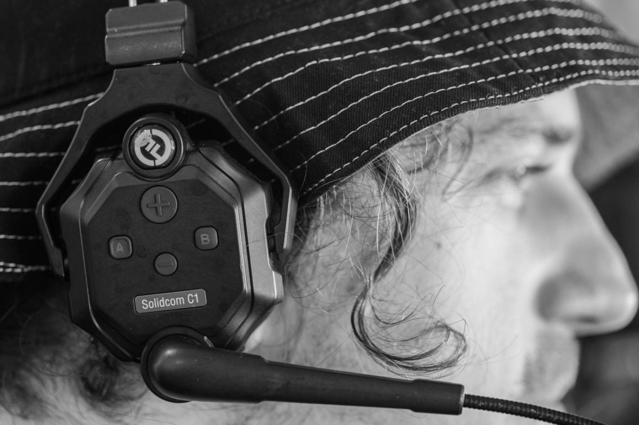 A black and white close-up of a person wearing a gaming headset with a microphone. The headset is labeled "Solidoom G1," and the person is wearing a striped bucket hat. Focus is on the headset, with part of the person's face visible in profile.