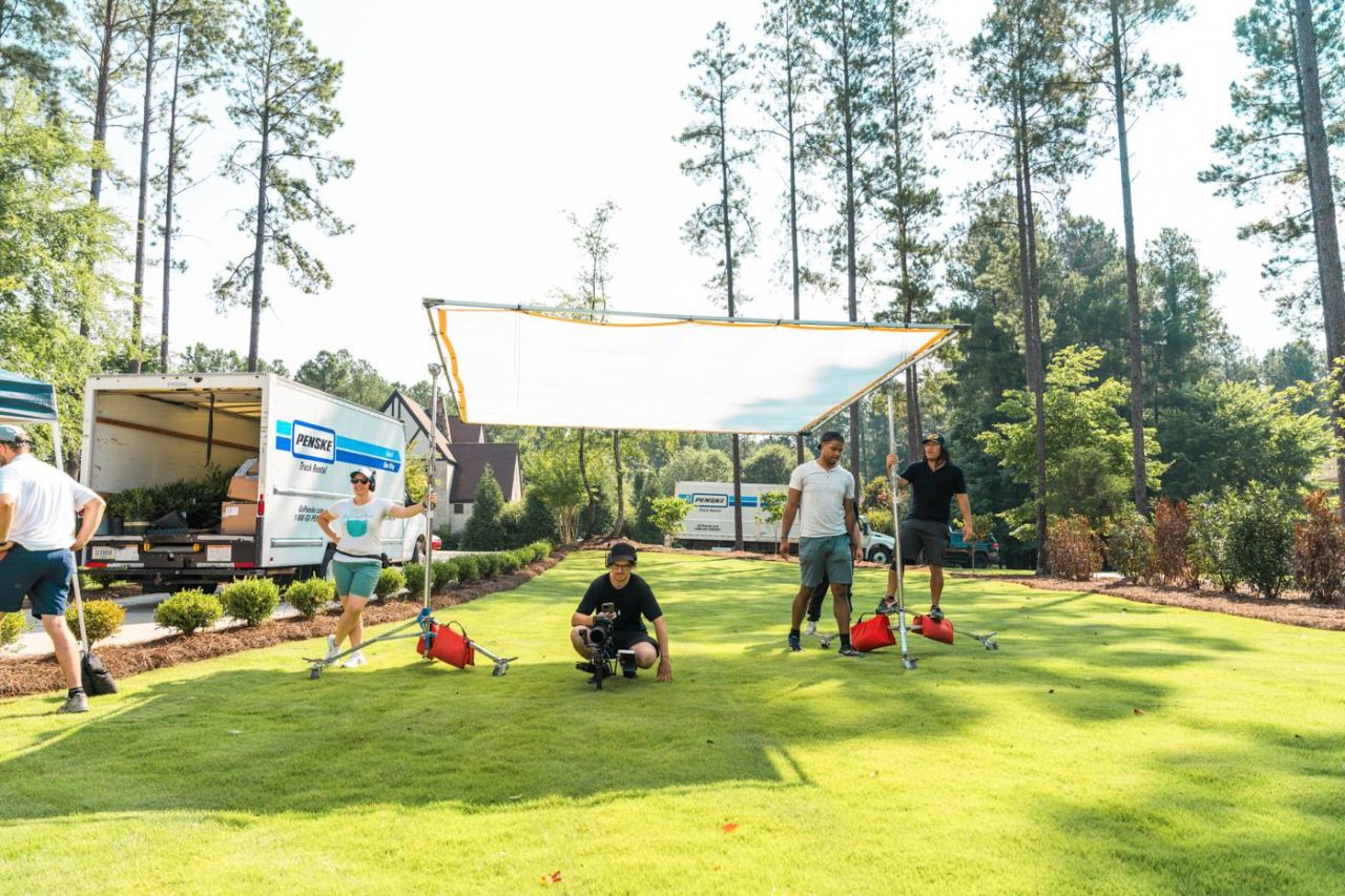 A group of people setting up a white canopy on a sunny lawn, surrounded by trees. Two trucks are parked nearby. Some individuals are holding equipment, while others adjust the canopy. The scene suggests an outdoor event preparation.