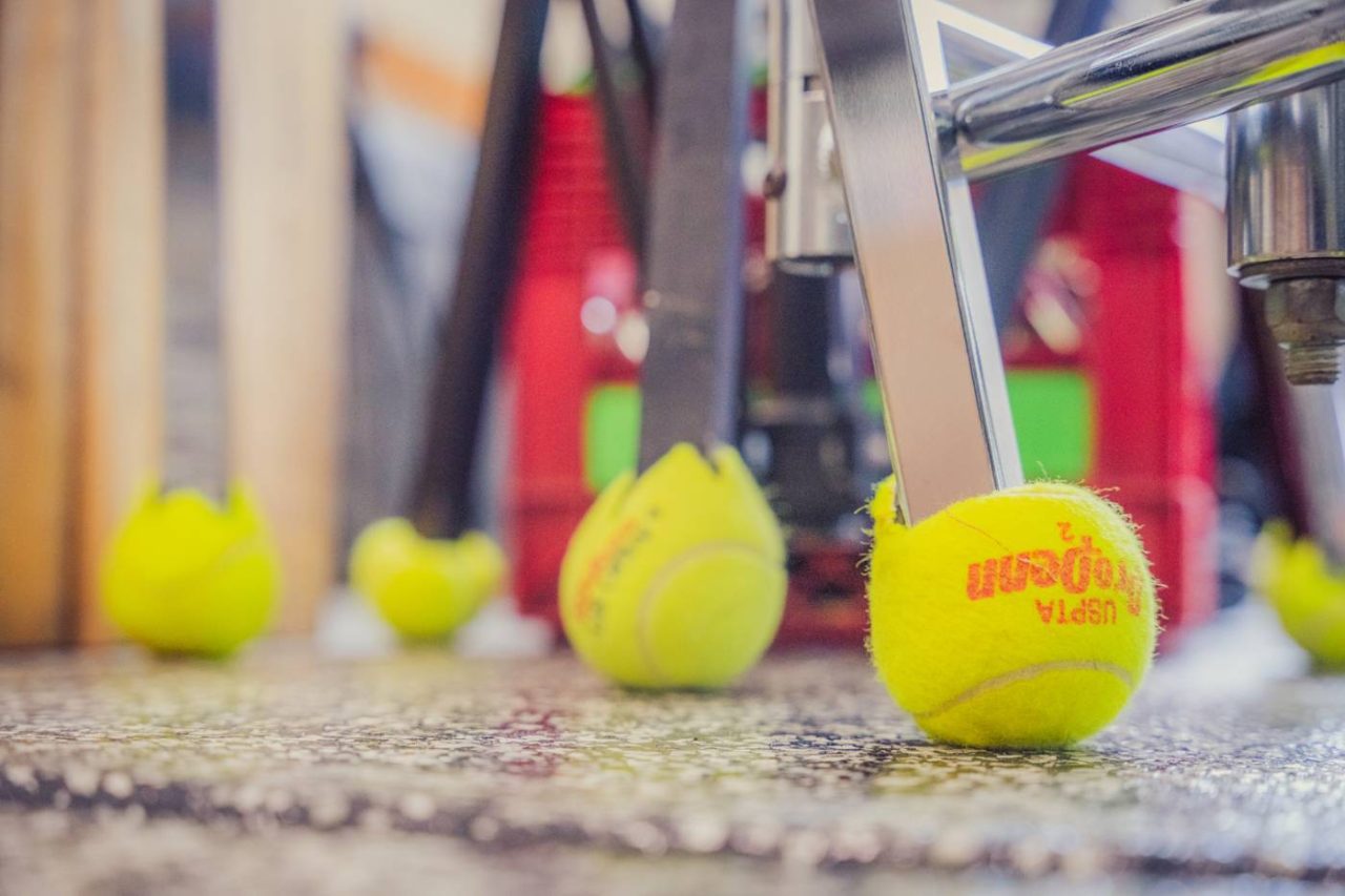 A close-up of a tennis ball used on the leg of a chair, with other tennis balls scattered on a speckled floor. A red crate is visible in the background.