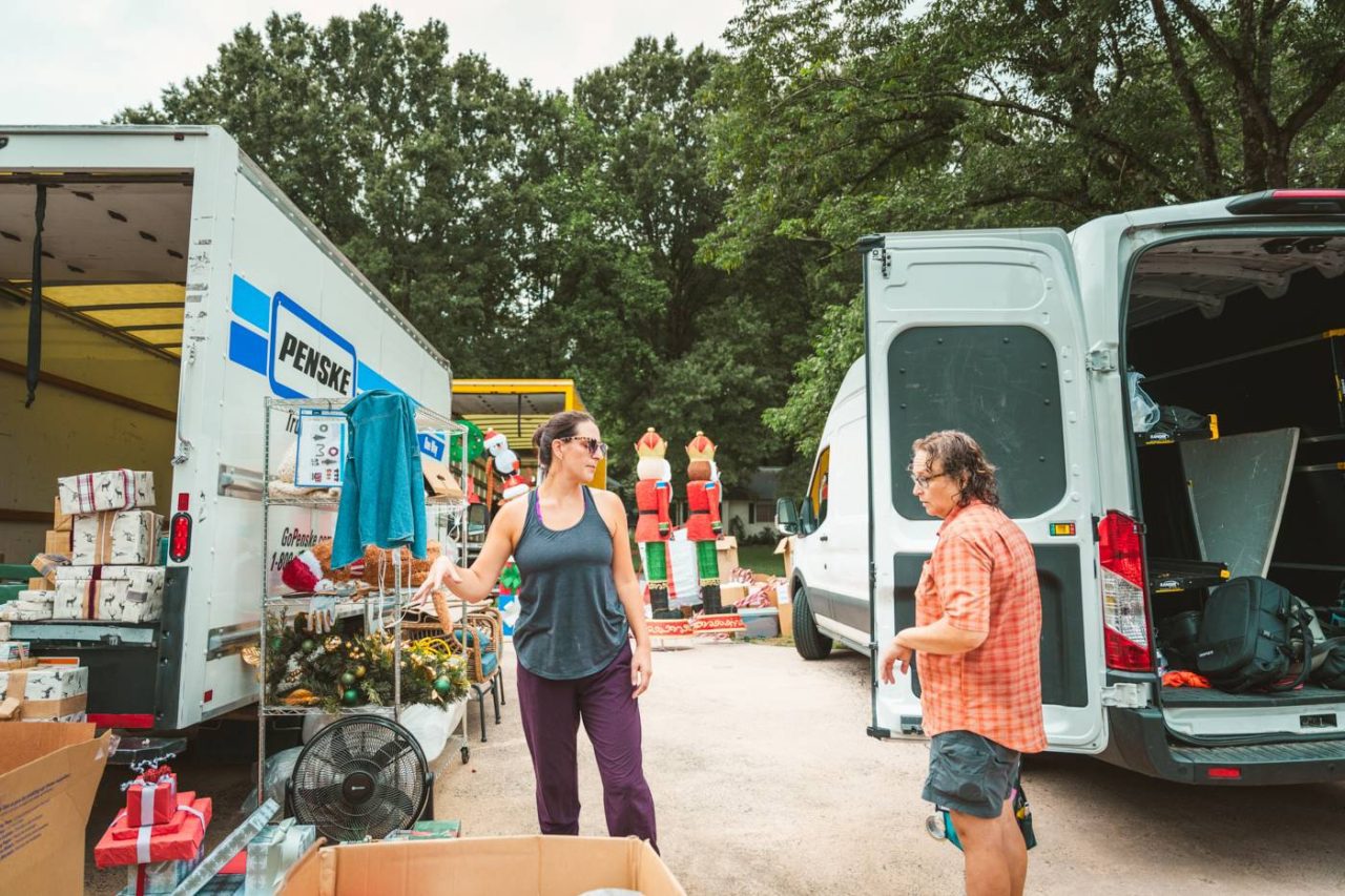 Two people stand between open moving trucks, surrounded by boxes and various items, including clothes and furniture. Trees are in the background, indicating an outdoor setting.