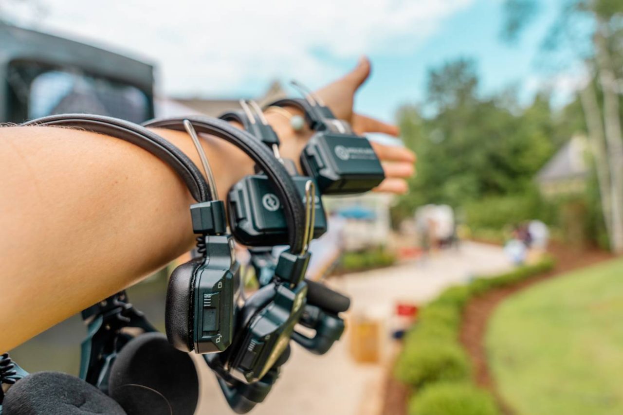 A person extends their arm, displaying several earpieces and communication devices looped around it. The background shows an outdoor setting with blurred trees and a few people in the distance.