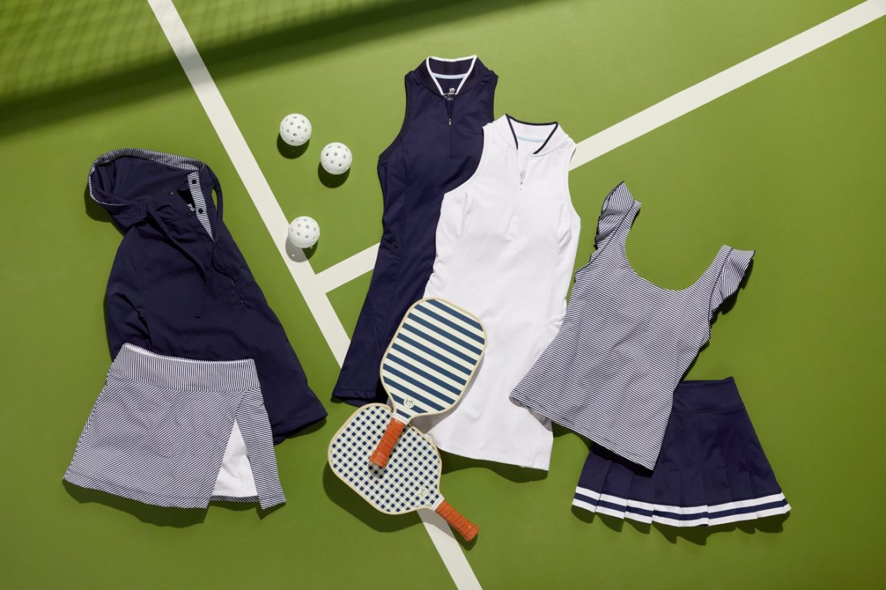 A collection of pickleball gear and clothing laid out on a green court. Includes a navy jacket, a white and navy sleeveless dress, a striped tank, a navy pleated skirt, a blue and white skort, paddles, and balls.