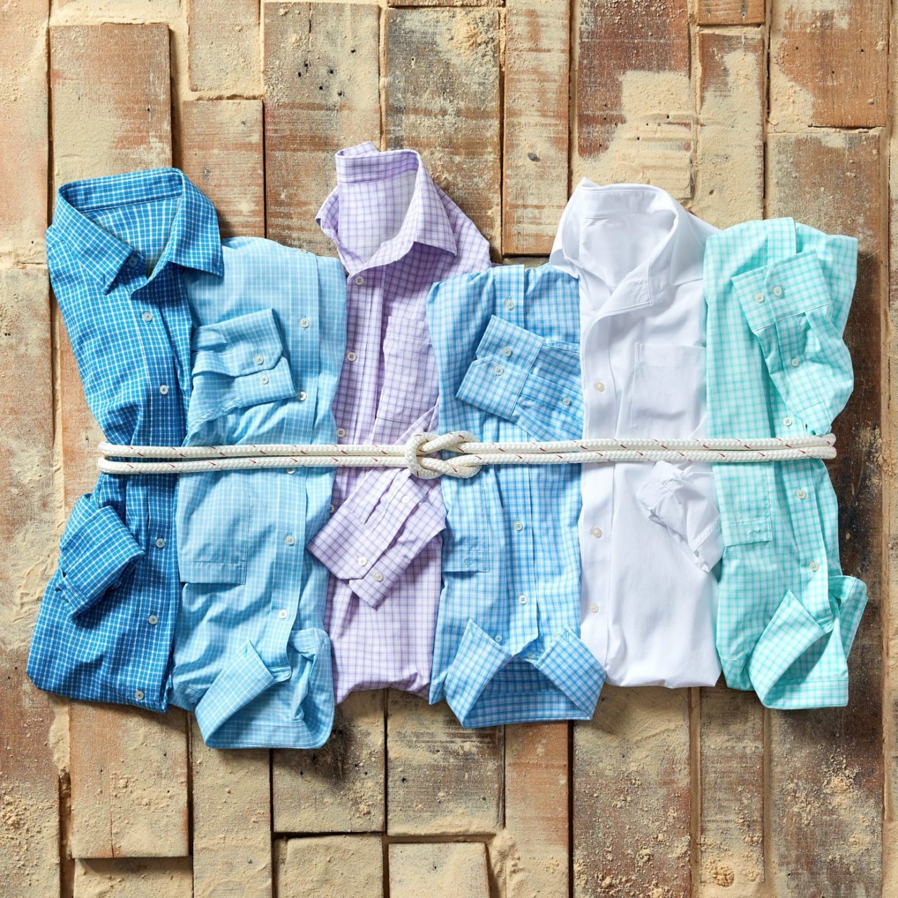 Five button-up shirts in shades of blue, purple, and white are neatly folded and tied together with a rope on a textured wooden surface.