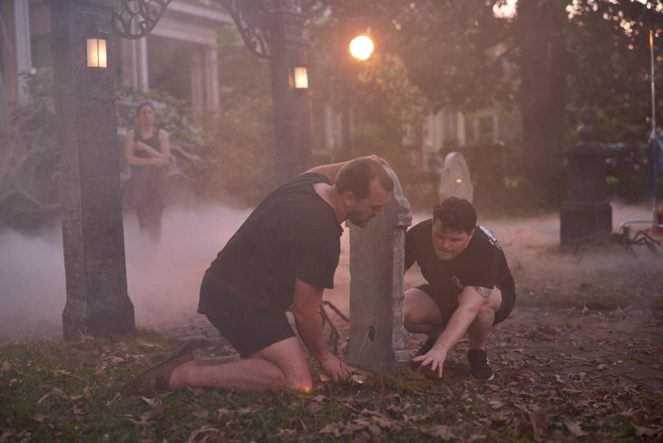 Two people kneeling by a gravestone in a misty, eerie setting, adjusting or positioning it. Soft, golden sunlight filters through surrounding trees. A third person stands watching in the background.