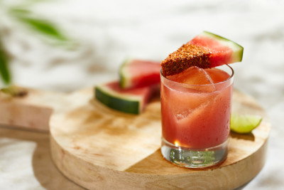 Natural light photo of a pink cocktail in a gleaming glass topped with a wedge of watermelon.