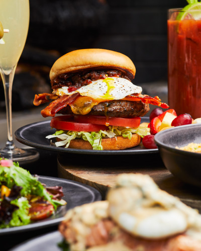 Low-angle food photography of a burger with toppings in a cluster of side dishes and delicious drinks.