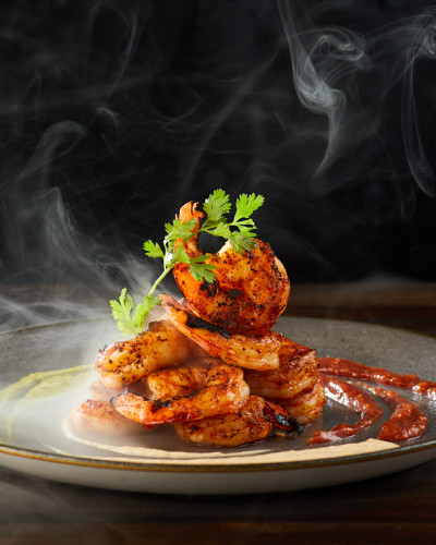 Dramatic on-black photograph of a plate of grilled shrimp with steam curling.