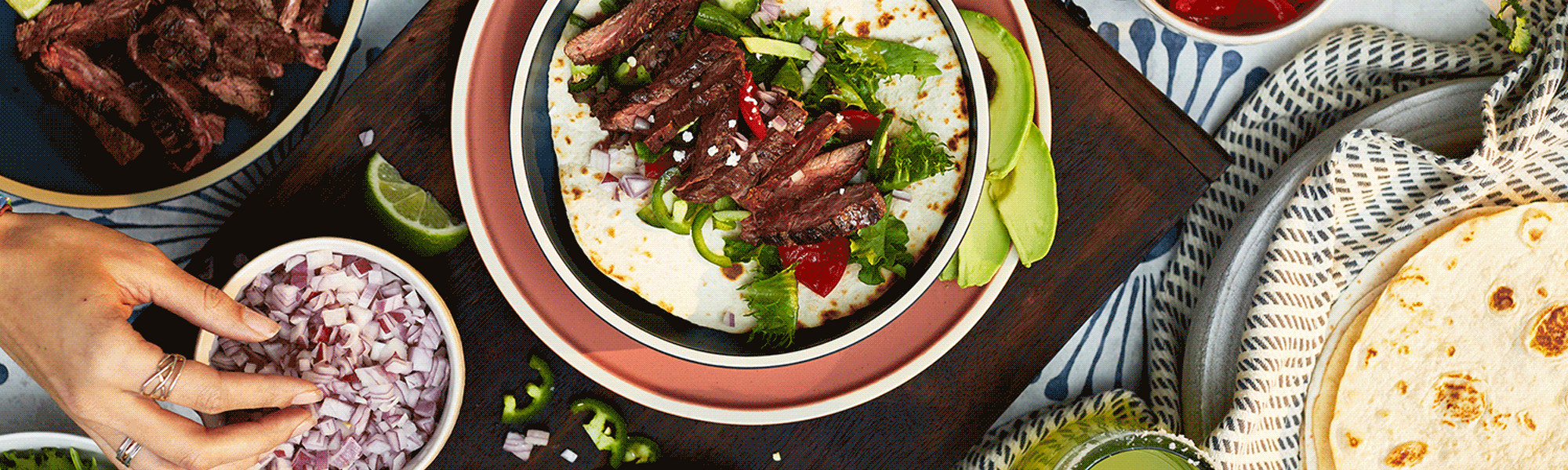 Photograph of colorful bowls of food from above with a hand assembling a steak taco.